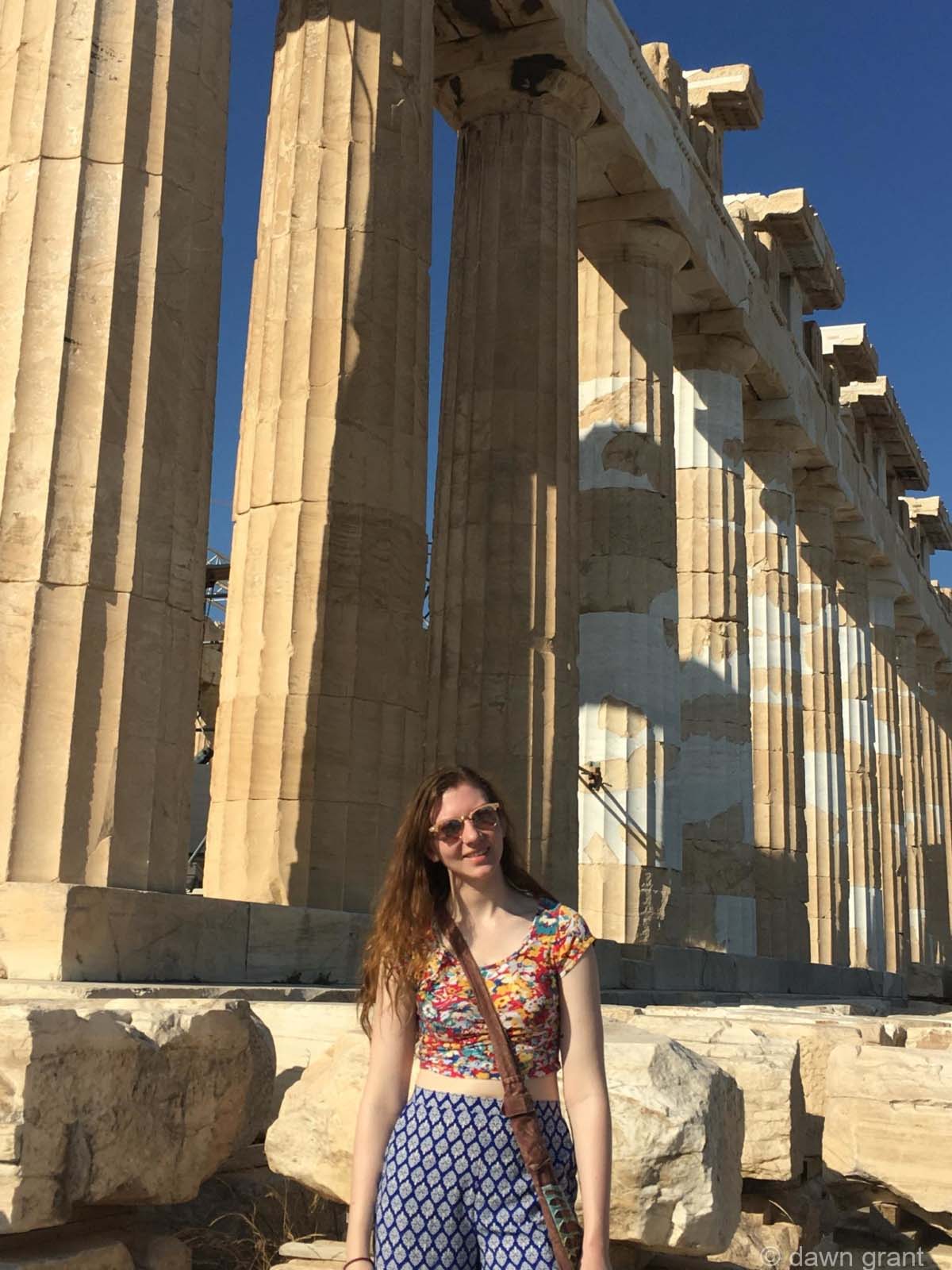 A young woman stands in front of the Parthenon ruins showing large columns that are obviously very old.