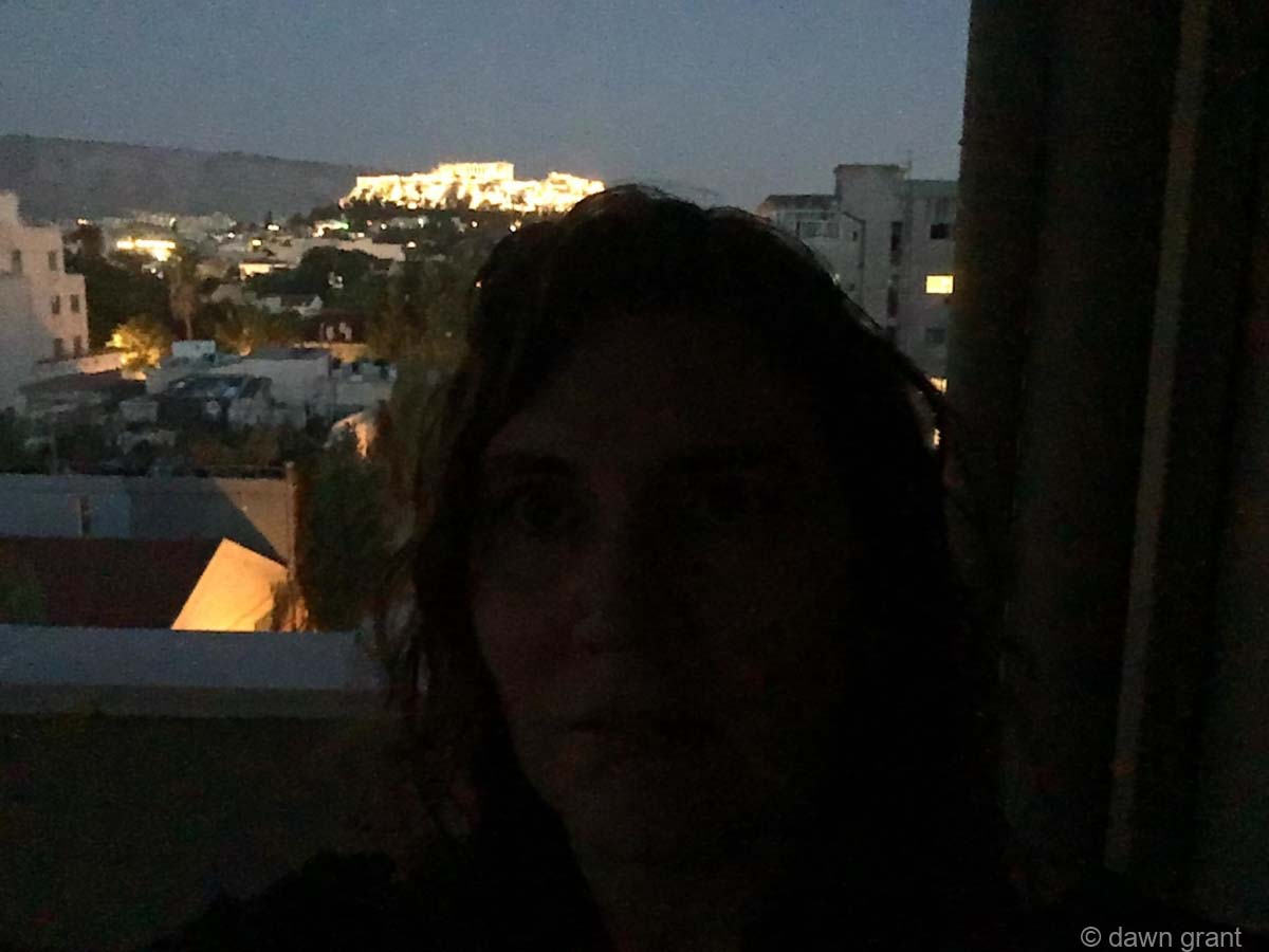 A photo taken at night with person in the foreground, city lights behind, and the Acropolis lit up on the hill.